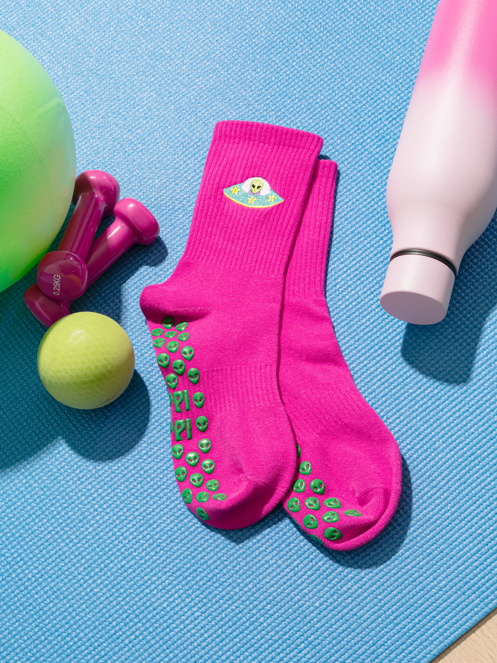 Pink socks with green alien grips, green ball, pink dumbbells, and green balloon on a blue mat. Slip-resistant socks for pilates, barre, lagree or yoga.