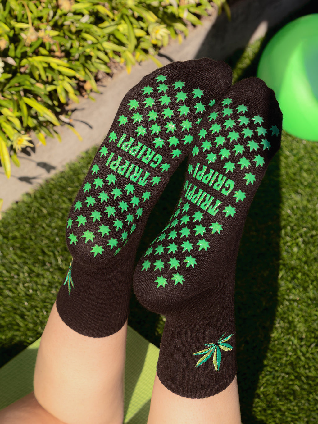 Black grip socks with green weed leaf patterns and 'TRIPPI GRIPPI' text on grass.  Bottom of stoner grip socks. Slip-resistant socks for pilates, barre, lagree or yoga.