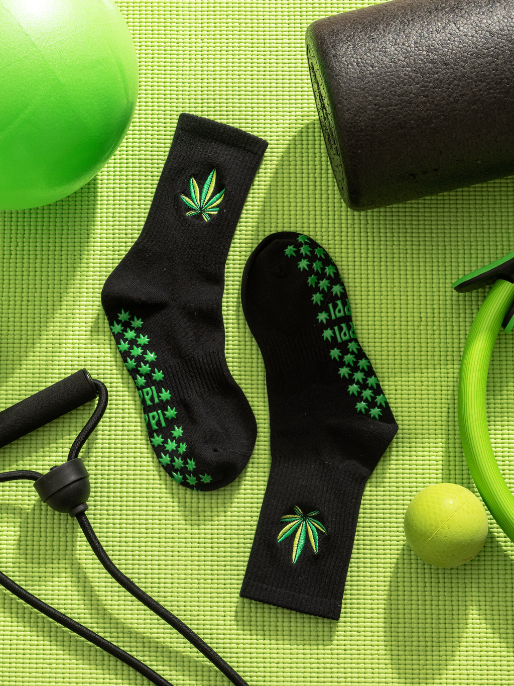 Black grip socks with green cannabis leaf patterns on a green mat with fitness equipment. Slip-resistant socks for pilates, barre, lagree or yoga.