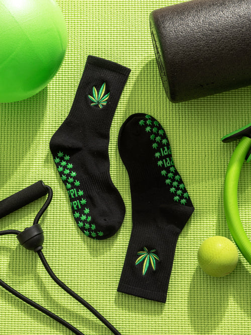 Black grip socks with green cannabis leaf patterns on a green mat with fitness equipment. Slip-resistant socks for pilates, barre, lagree or yoga.